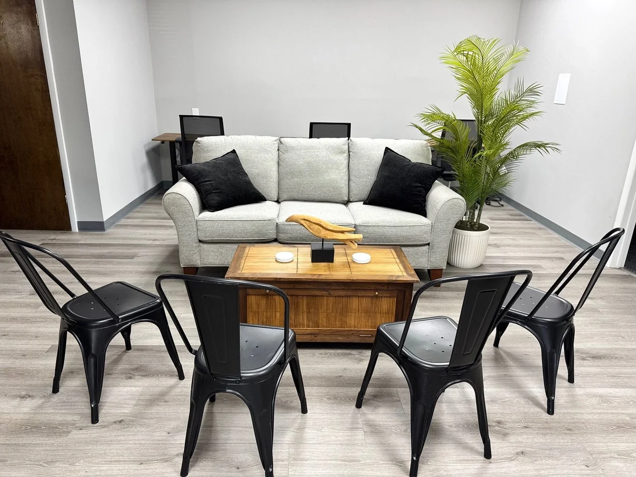 A modern living room with a light-colored upholstered sofa, four black metal chairs, a wooden coffee table, a potted green plant, and a small wooden desk with two chairs in the background.