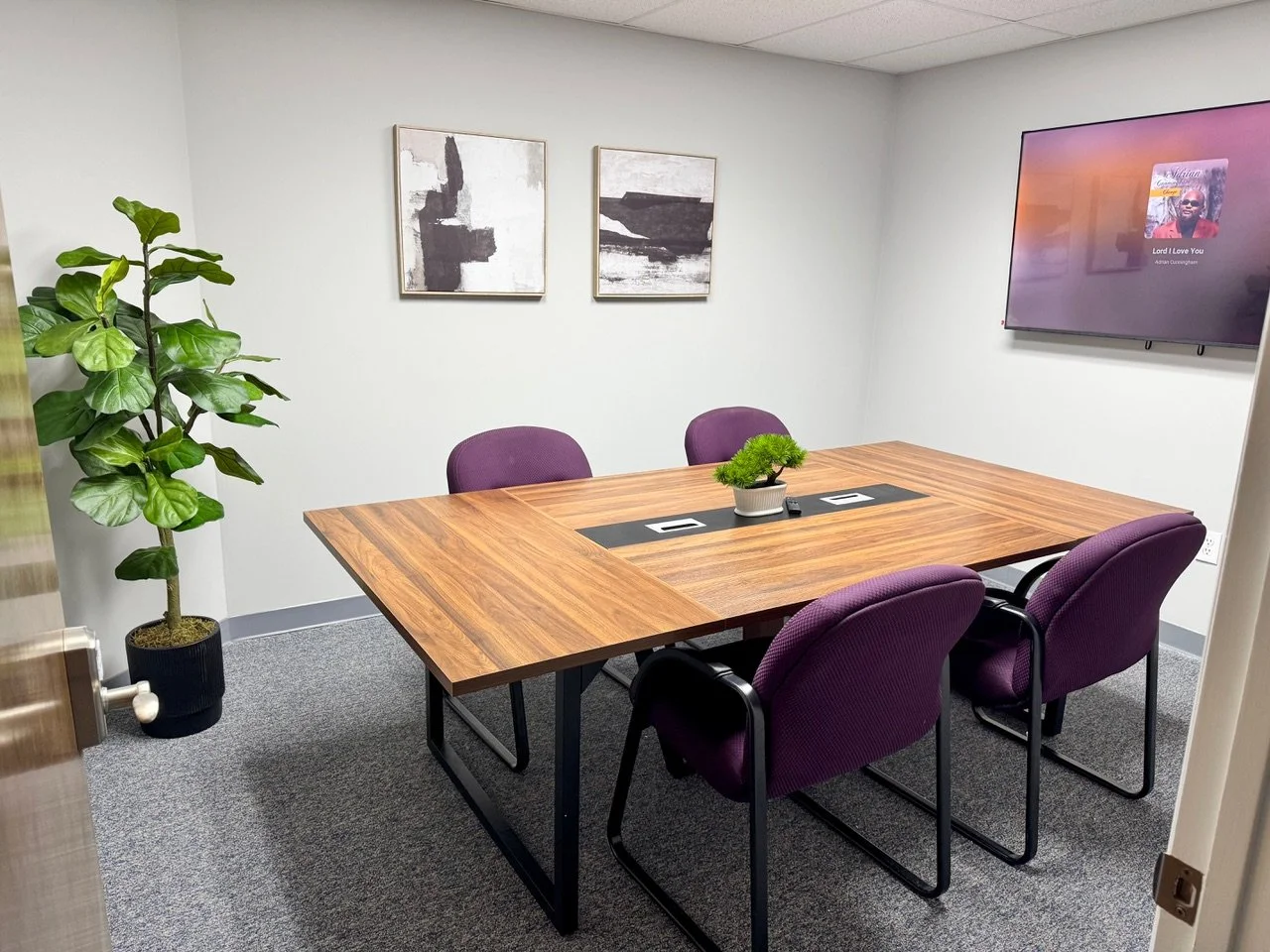 Empty conference room with wooden table, four purple chairs, a potted plant in the center, a blank wall with two abstract paintings, and a large wall-mounted TV.