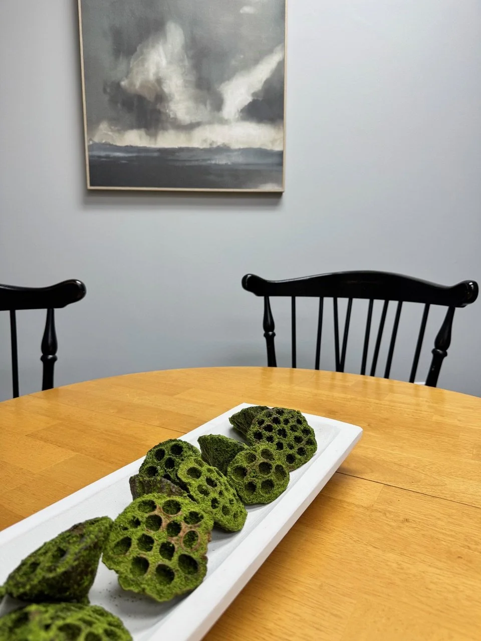 Close-up of a white rectangular platter with slices of green lotus root on a wooden table. In the background, there are two black wooden chairs and a framed abstract painting on a light blue wall.