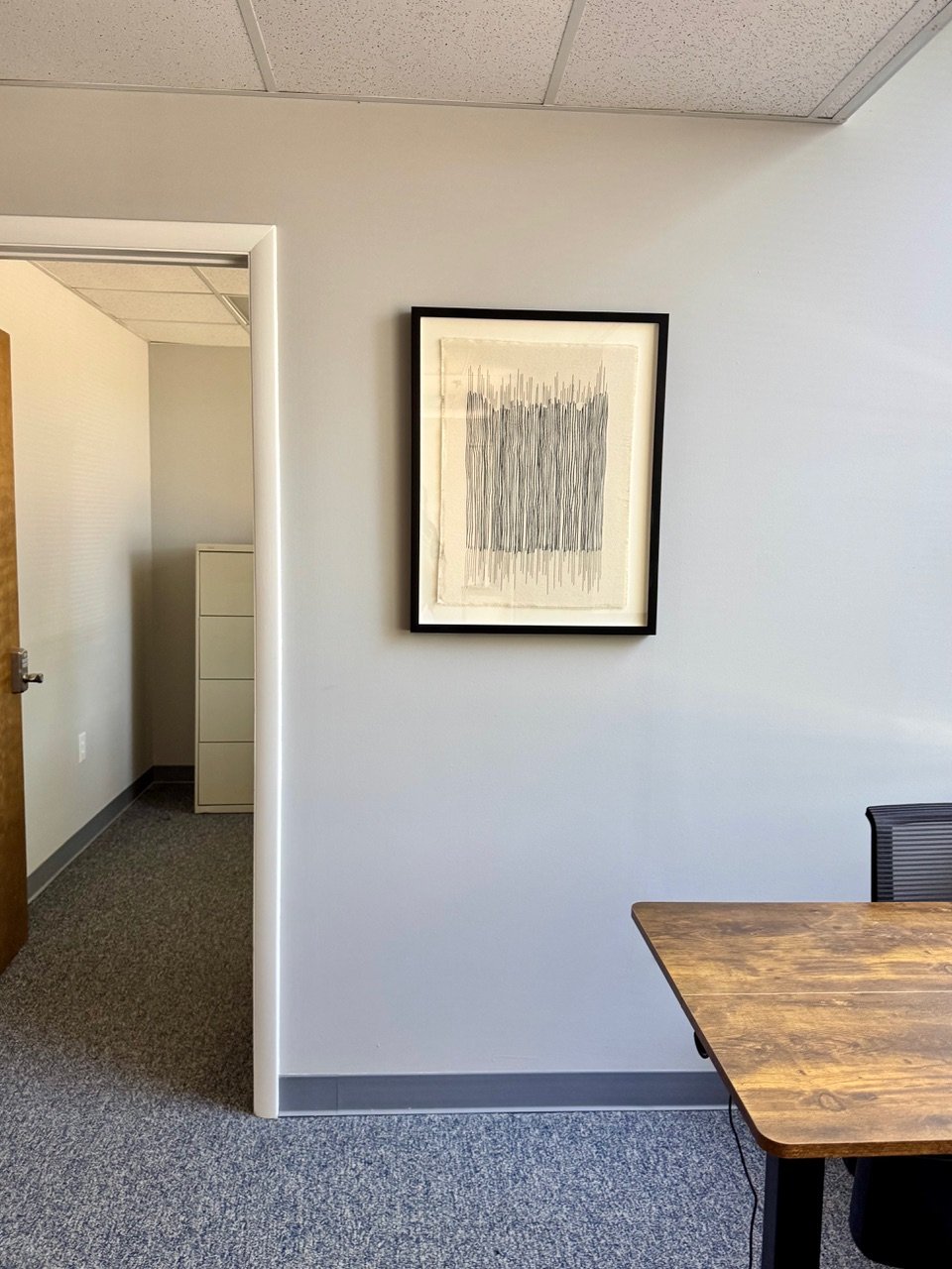 An office room with a framed abstract black and white artwork on the wall, a wooden table partially visible, and a doorway leading to a small hallway with a white filing cabinet.