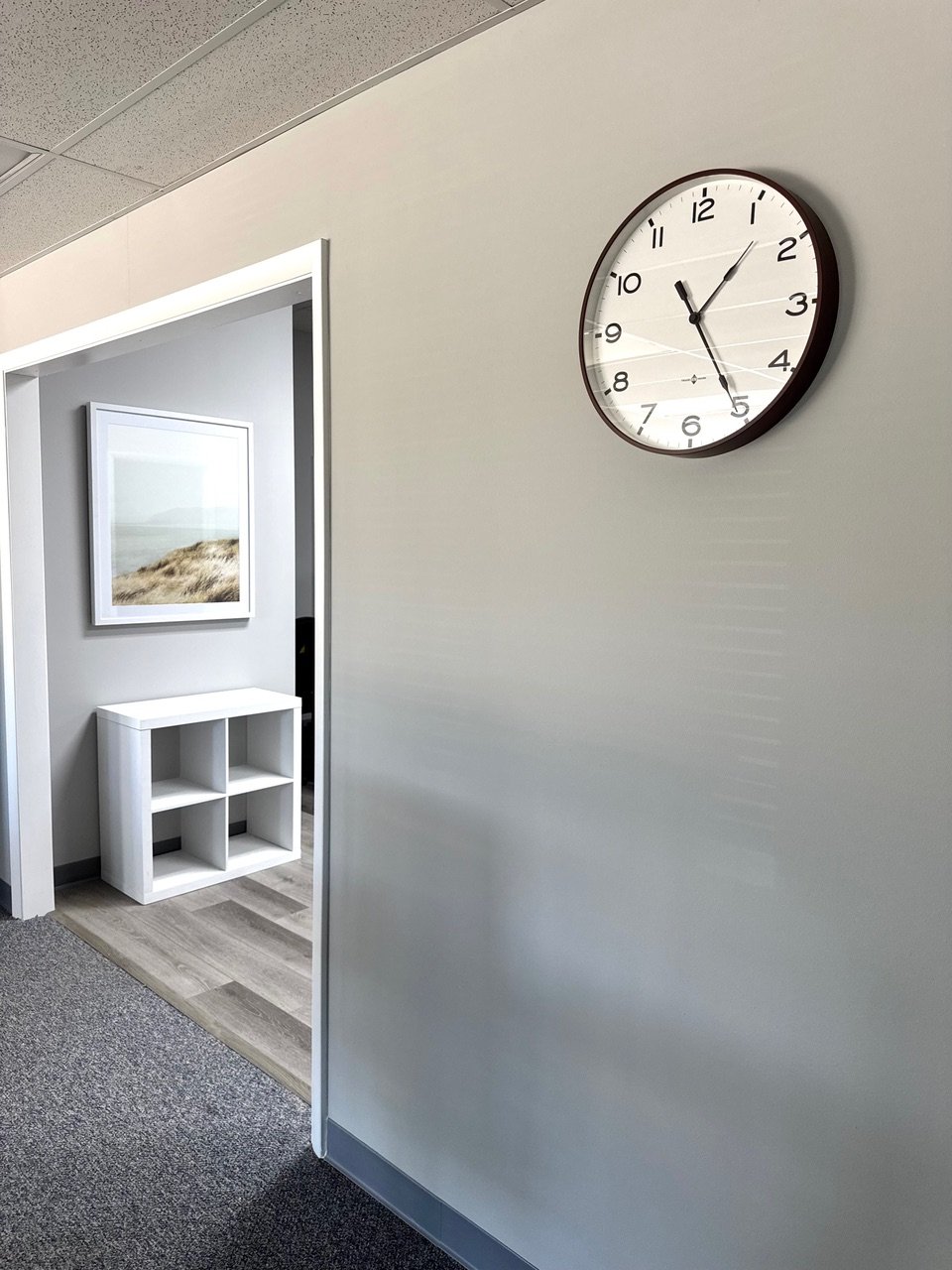 Office hallway with a round wall clock showing 1:24, a white cubical shelf, and a framed picture of sand dunes.