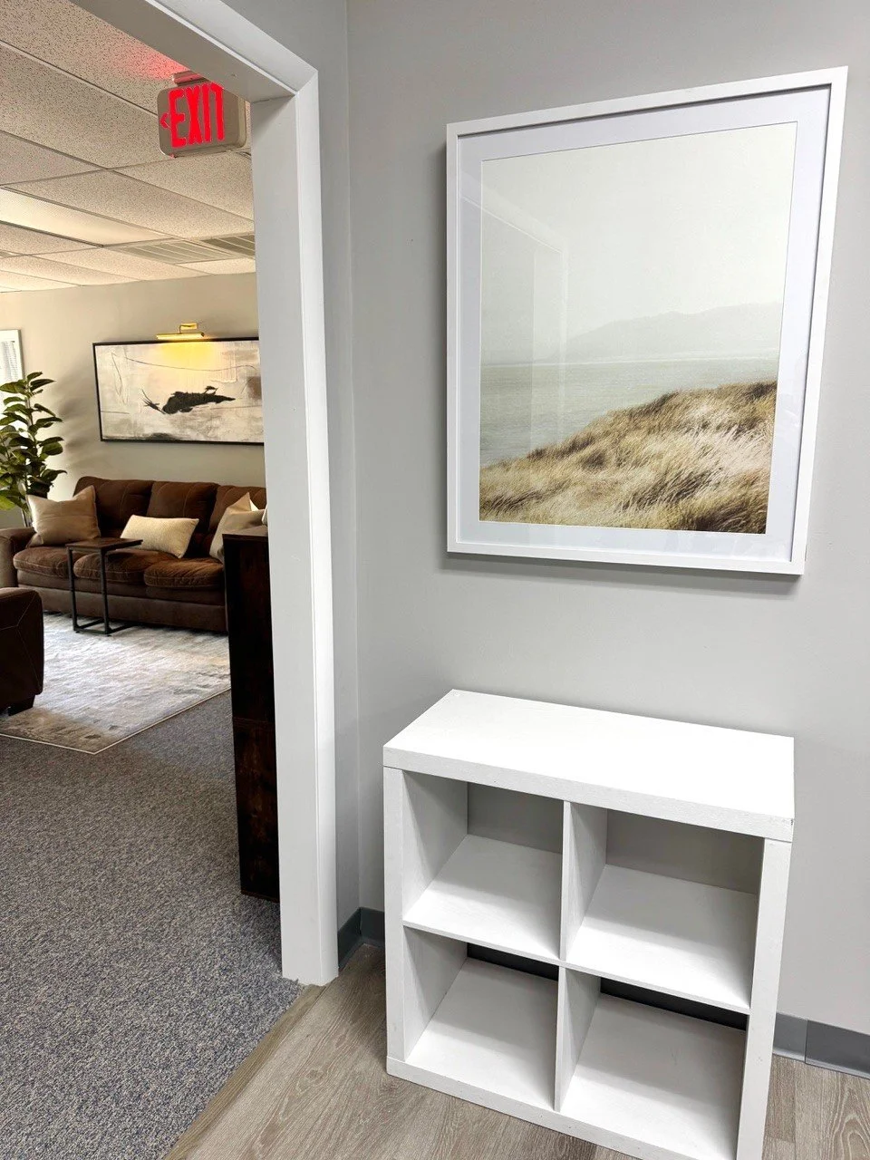 Interior view of a waiting area or office with a white shelf, a framed beachscape photo on the wall, and part of a lounge with sofa and pillows visible in the background.