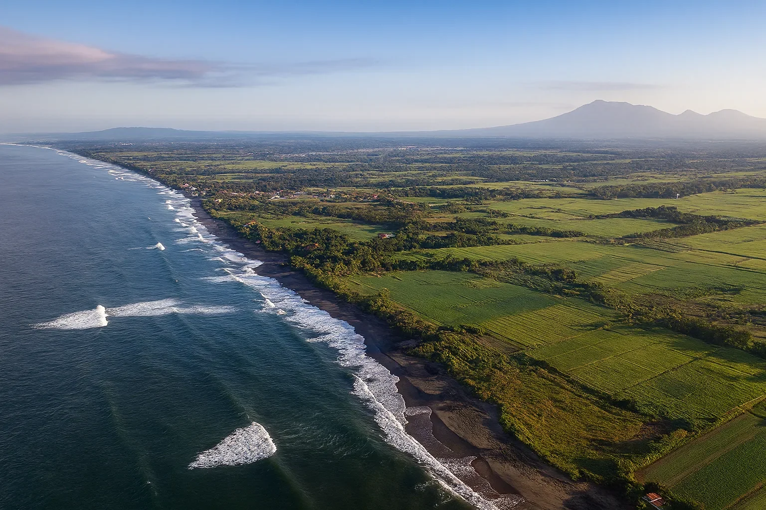 Aerial view of a coastline with waves crashing on the beach, surrounded by green fields and mountains in the background during daytime.