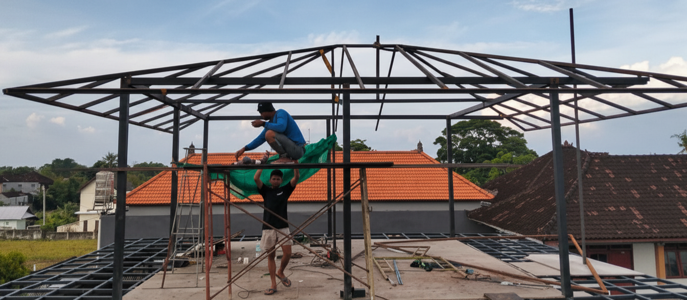 Two construction workers are building a metal framework on a rooftop, with one worker standing underneath and another on top, working on the structure. The background shows houses with orange and dark roofs under a partly cloudy sky.