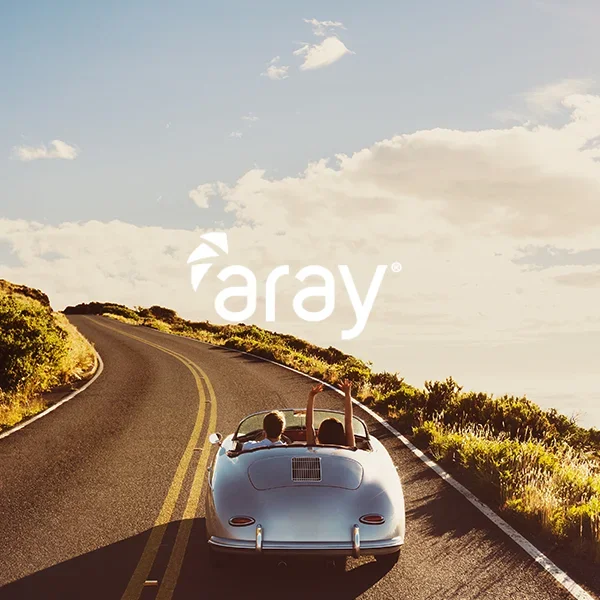A white convertible car driving on a winding coastal road with green bushes on the sides, under a partly cloudy sky. The driver and passenger are enjoying the ride, with one person waving.