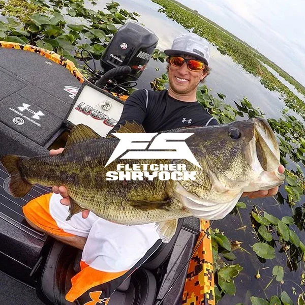 A man on a boat in a lake holding a large bass fish, smiling at the camera, with water and lily pads in the background.