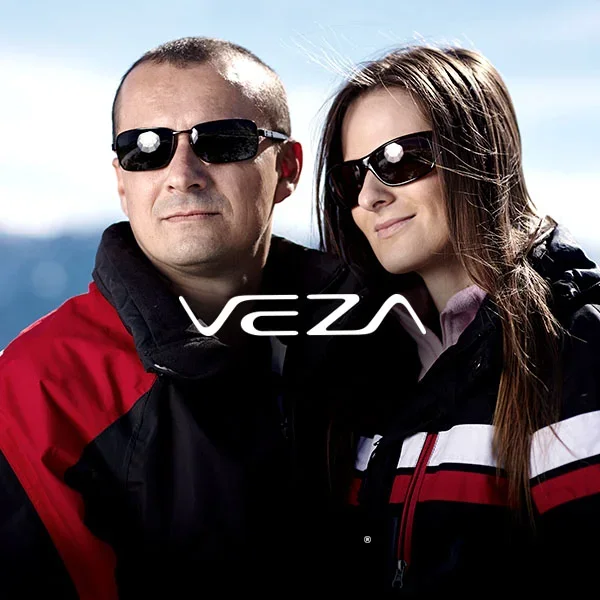 A man and woman wearing sunglasses and outdoor winter jackets, standing together outdoors with a mountain landscape in the background.
