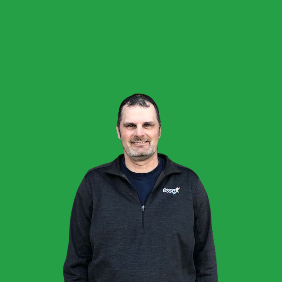 A man with dark hair and a beard smiling, wearing a black jacket with an 'essex' logo, standing against a green background.