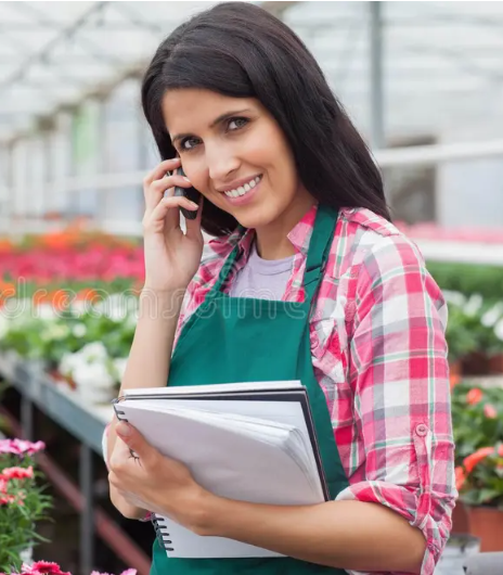 A smiling woman with dark hair in a greenhouse, wearing a plaid shirt and green apron, holding a clipboard and talking on the phone.