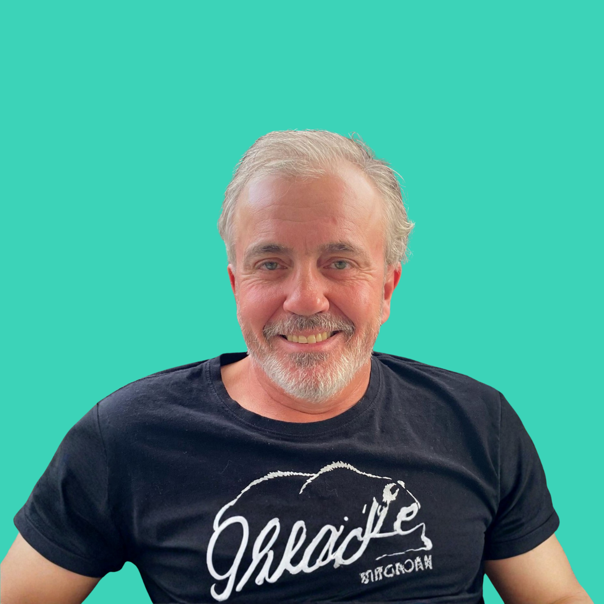 A smiling man with gray hair and beard, wearing a black t-shirt with a chocolate logo, against a teal background.