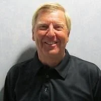 A middle-aged man with blonde hair smiling, wearing a black shirt, standing against a plain gray background.