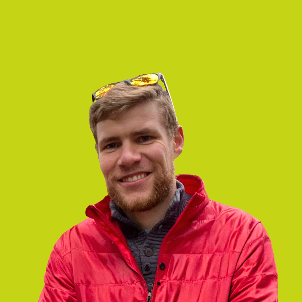 A young man with short brown hair and a beard, smiling, wearing a red jacket and sunglasses on his head, with a green background.