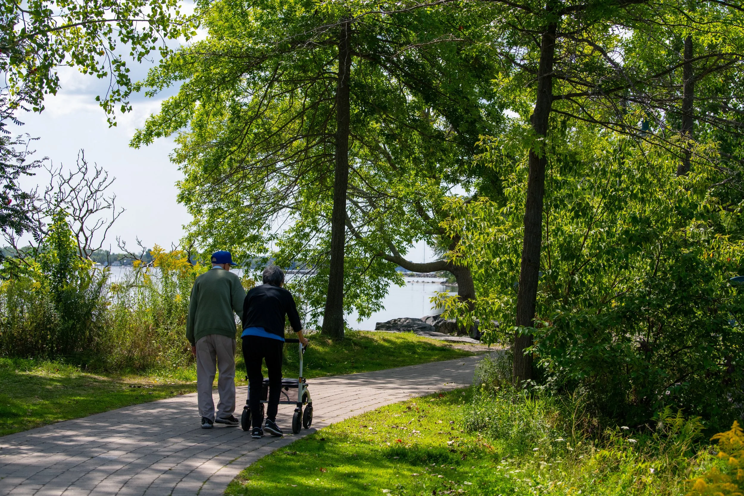 Two elderly people walking with a walker on a paved path through a park with lush green trees and a body of water in the background.