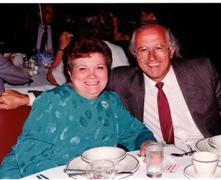 A smiling middle-aged woman and an older man sitting at a table during a formal event, surrounded by dinnerware and glasses.