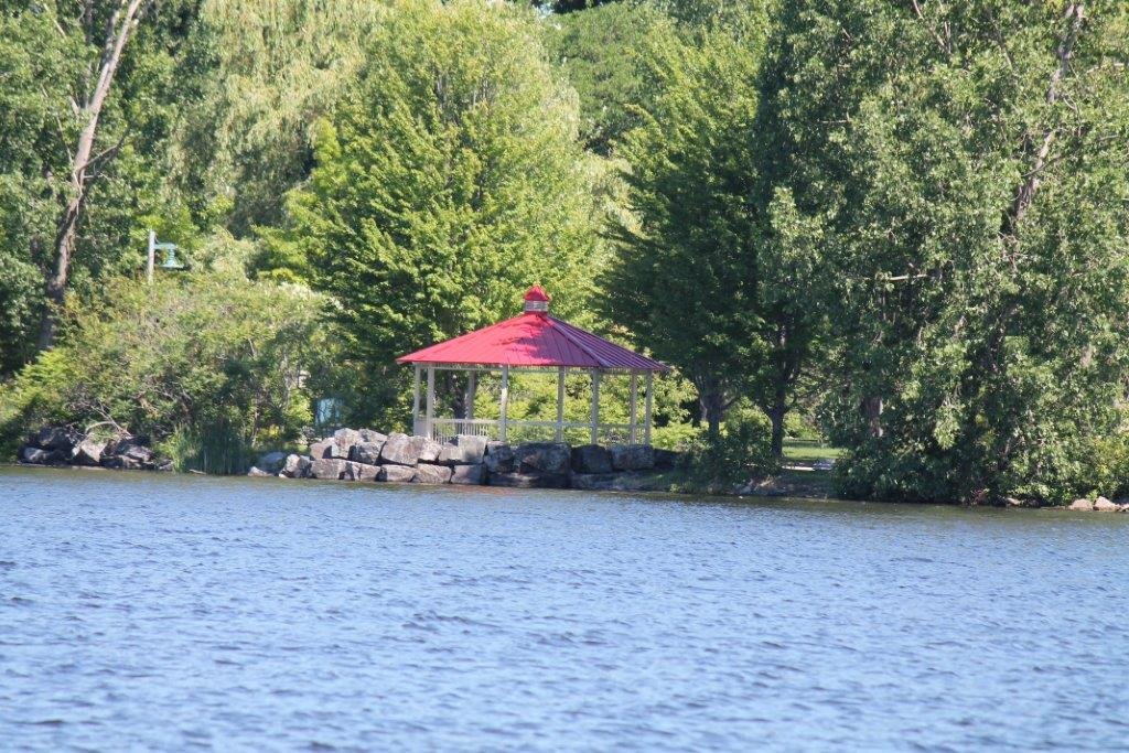 A lakeside scene with a small gazebo featuring a red roof, situated on rocks and surrounded by lush green trees.