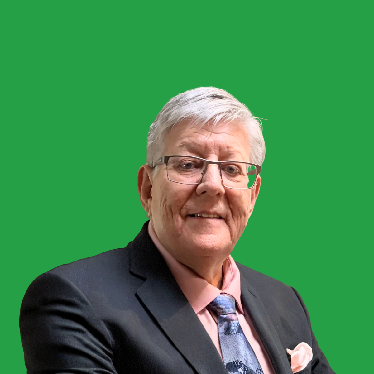 An elderly man with glasses and gray hair, wearing a black suit jacket, pink shirt, and a colorful tie, smiling against a plain green background.