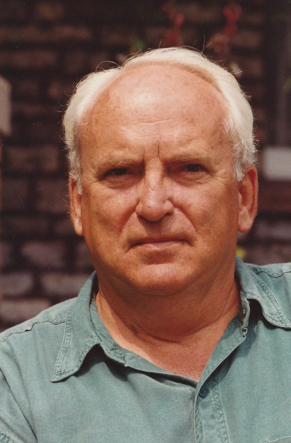 Close-up of an elderly man with white hair, wearing a light green collared shirt, standing outdoors with a blurred brick background.