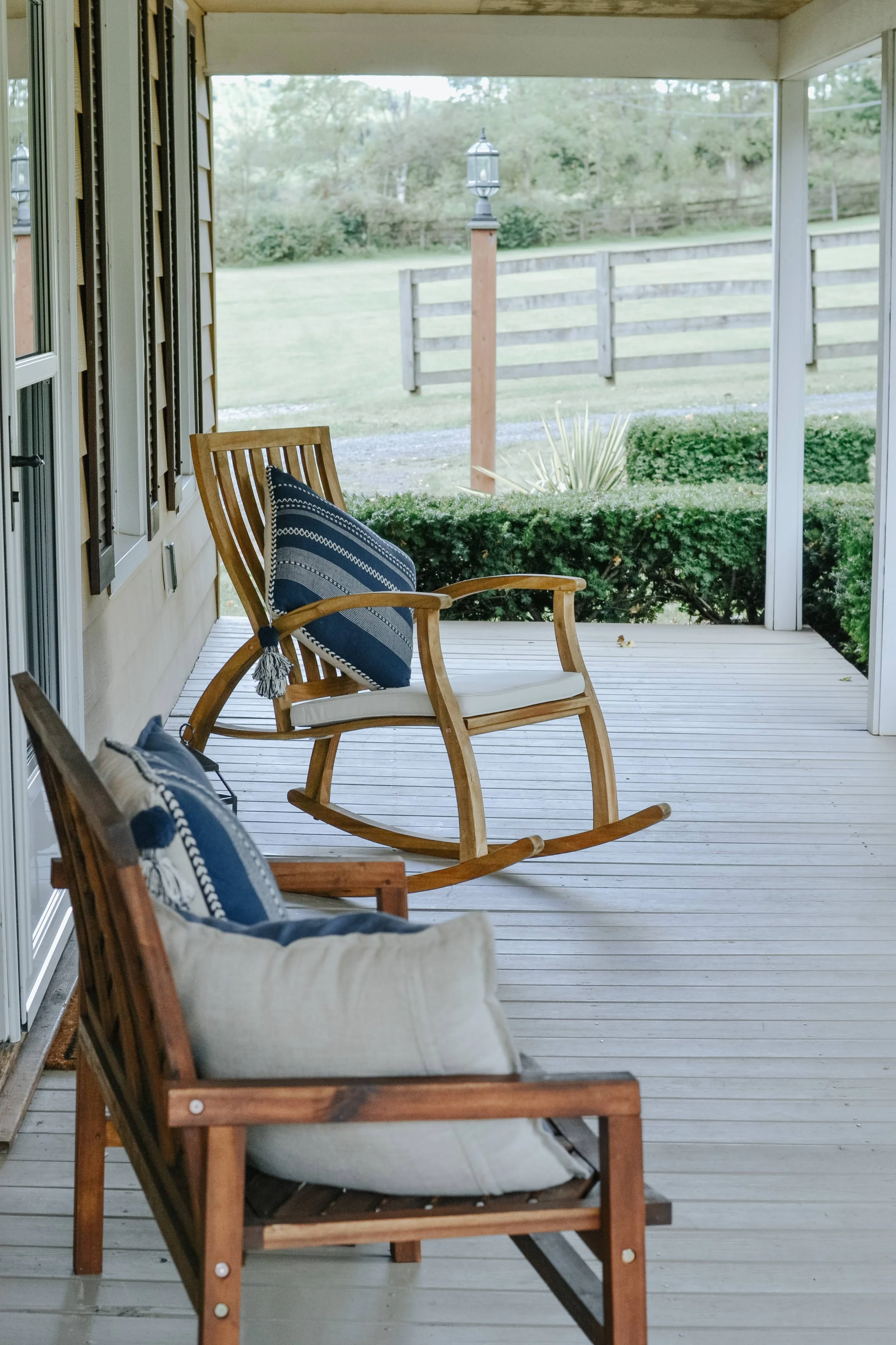 View of a covered porch with two wooden rocking chairs and a cushioned wooden outdoor chair, decorated with navy-blue pillows, overlooking a backyard with green grass, bushes, and a wooden fence in the distance.