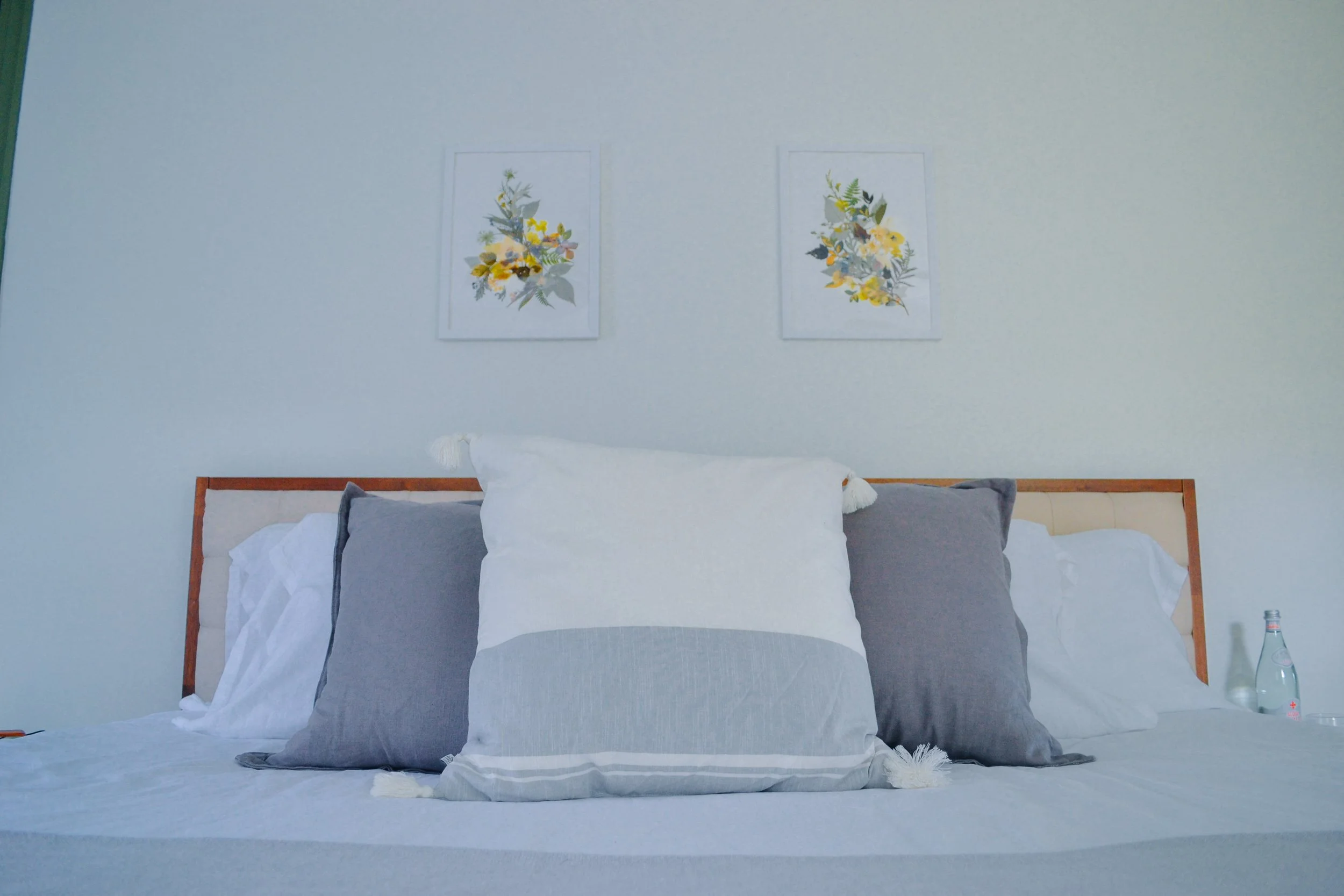 A neatly made bed with a wooden headboard, white and gray pillows, and a white duvet. Two framed floral art prints hang on the white wall above the bed. A glass water bottle and a glass are on the right side of the bed.