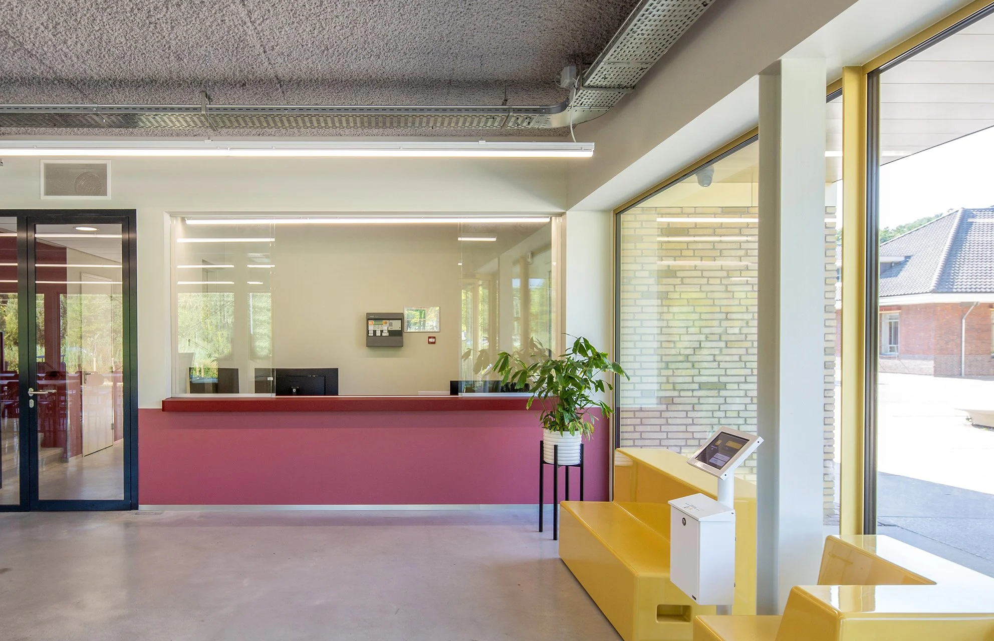 Binnenzijde van een modern gebouw met een balie, planten, grote ramen en zitplaatsen.