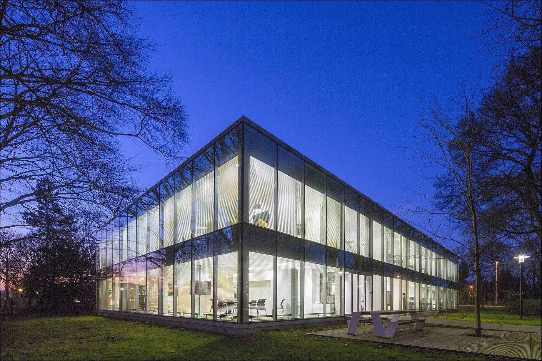 Moderne kantoorgebouw met veel ramen, verlicht van binnen, omgeven door bomen en gras, 's avonds.