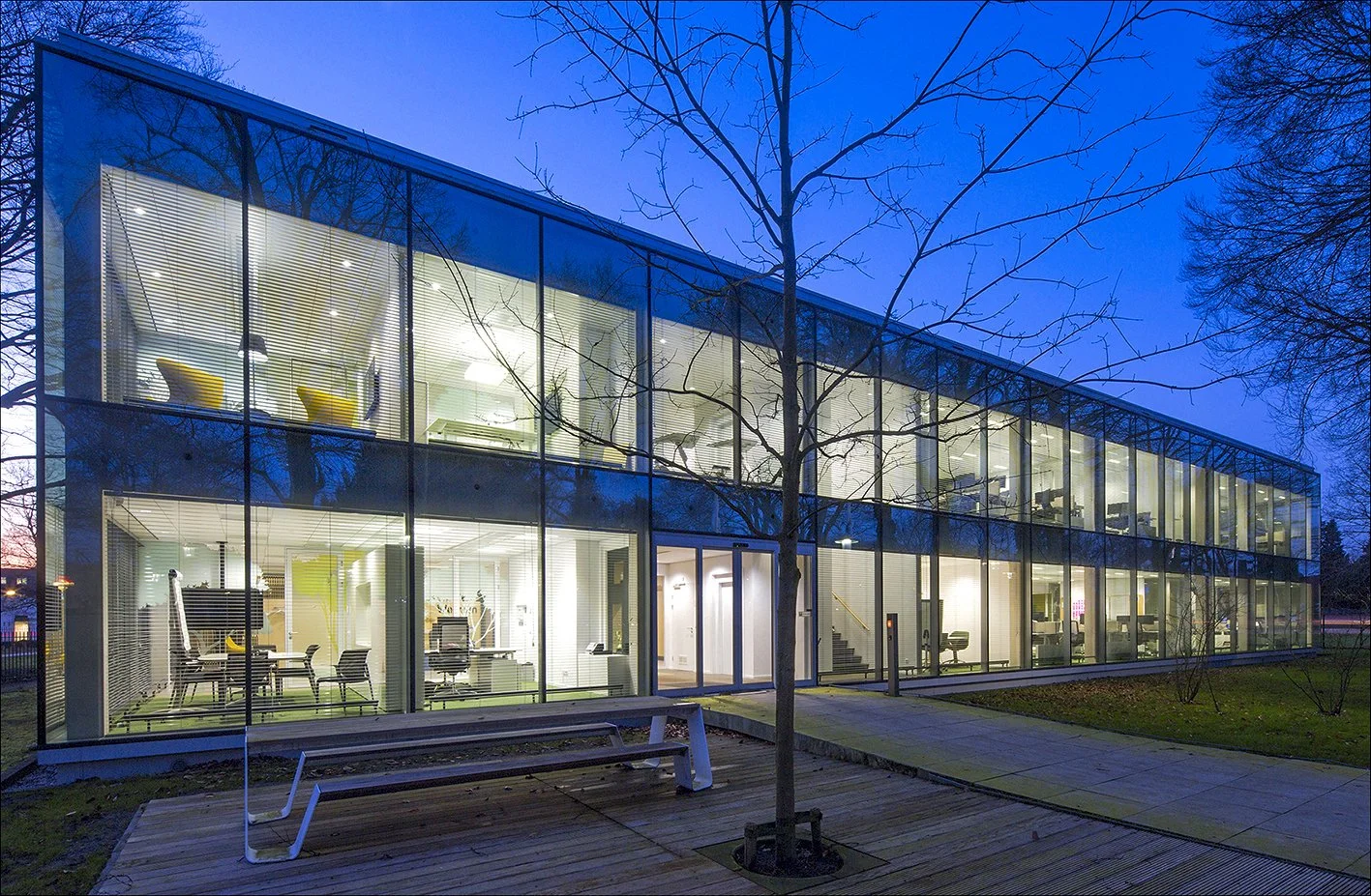 Een modern kantoorgebouw met glazen gevels, verlicht 's avonds, omringd door bomen zonder bladeren.