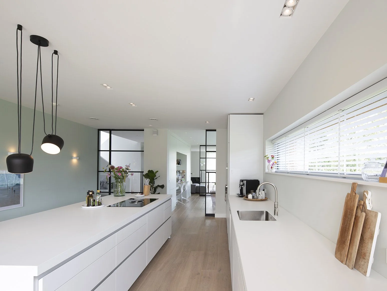 Moderne, lichte keuken met wit werkblad en eiland, hanglampen, grote ramen met jaloezien, planten, en een open indeling naar een eetkamer en woonkamer.