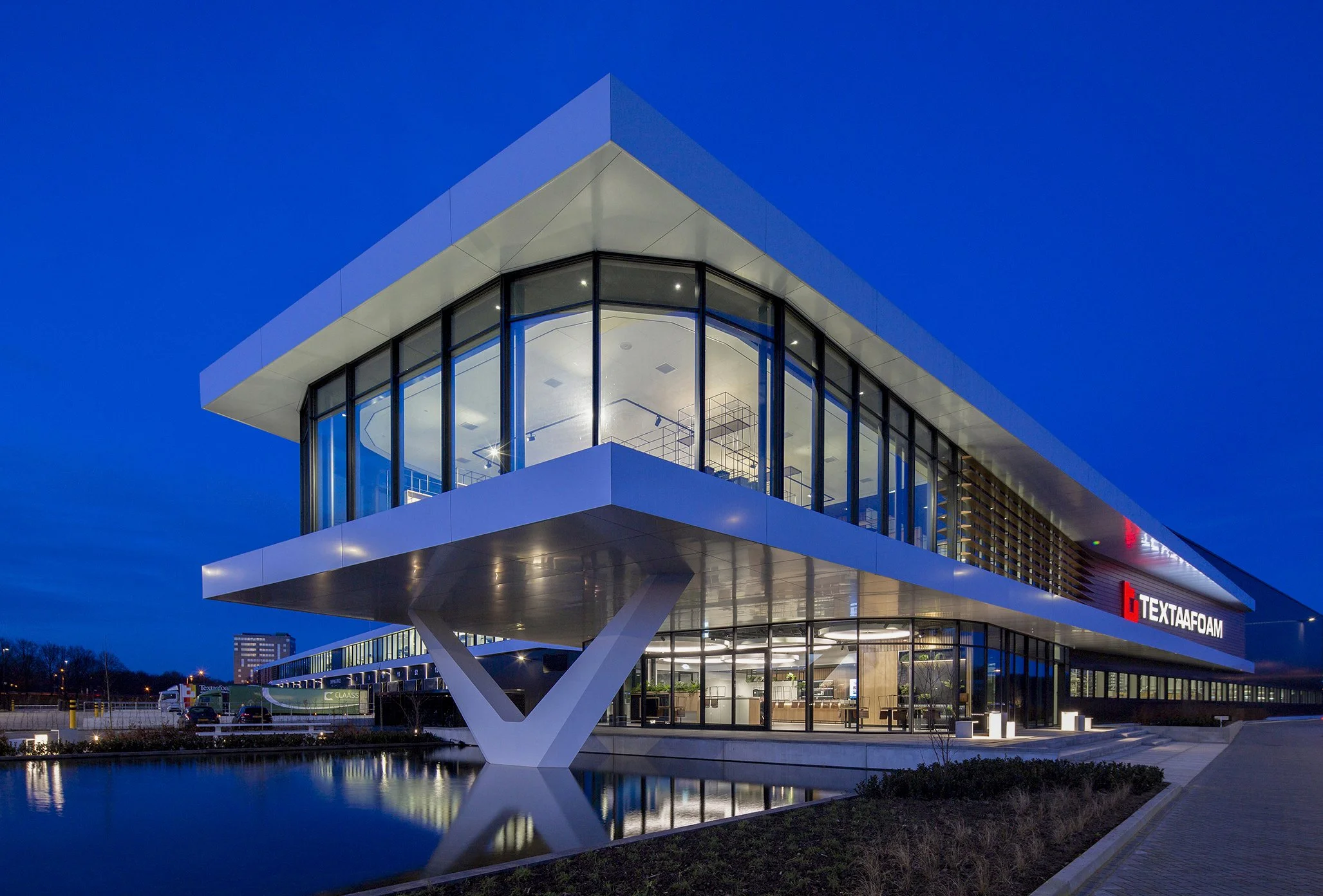 Een modern kantoorgebouw met grote ramen, verlicht van binnen, tegen een heldere blauwe avondlucht. Het gebouw staat boven een waterpartij die hetReflecteert.
