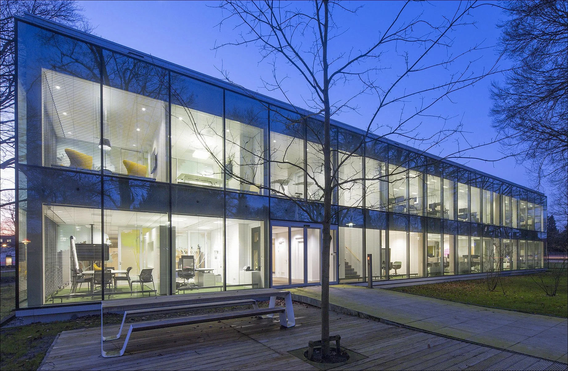 Modern glazen kantoorgebouw met verlichte binnenkant, omgeven door kale bomen en een houten bank in de tuin, bij schemering.