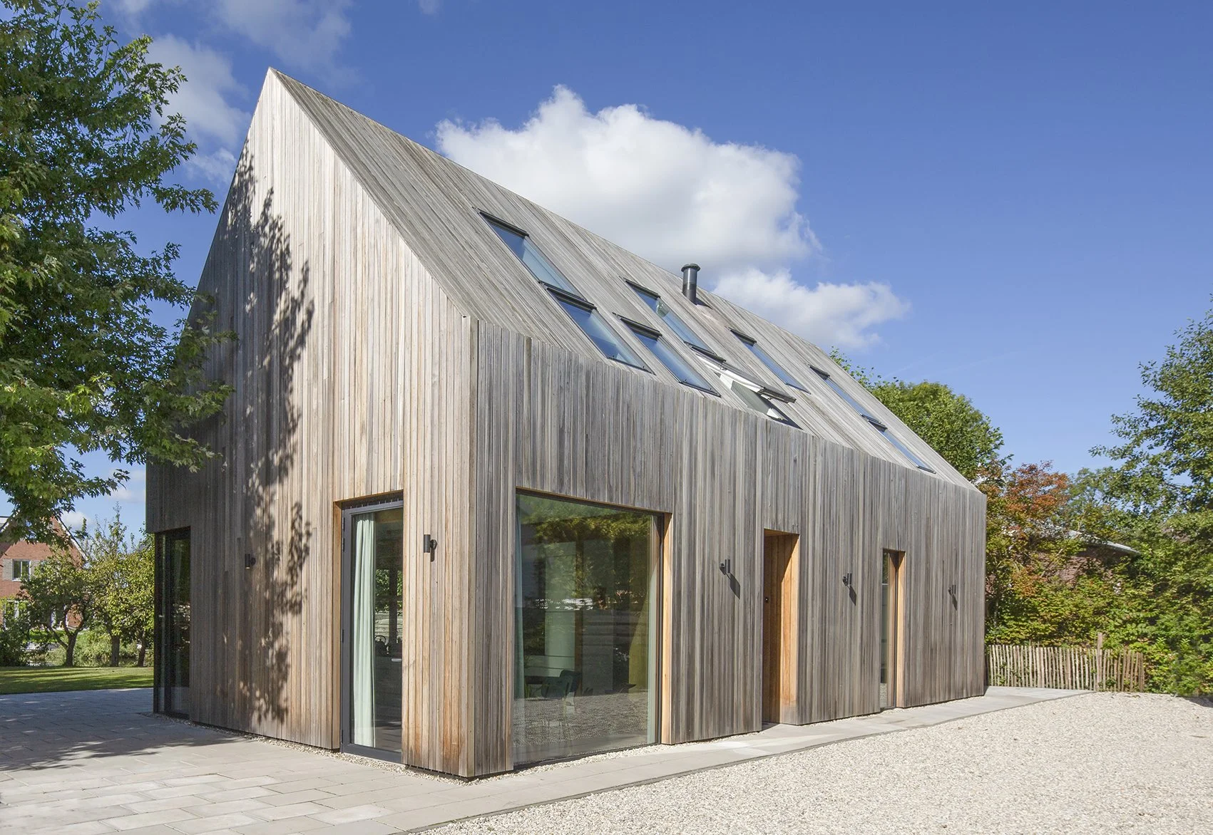 Houten modern huis met grote ramen en schuine daken onder een blauwe hemel met wolken, omgeven door groene bomen.