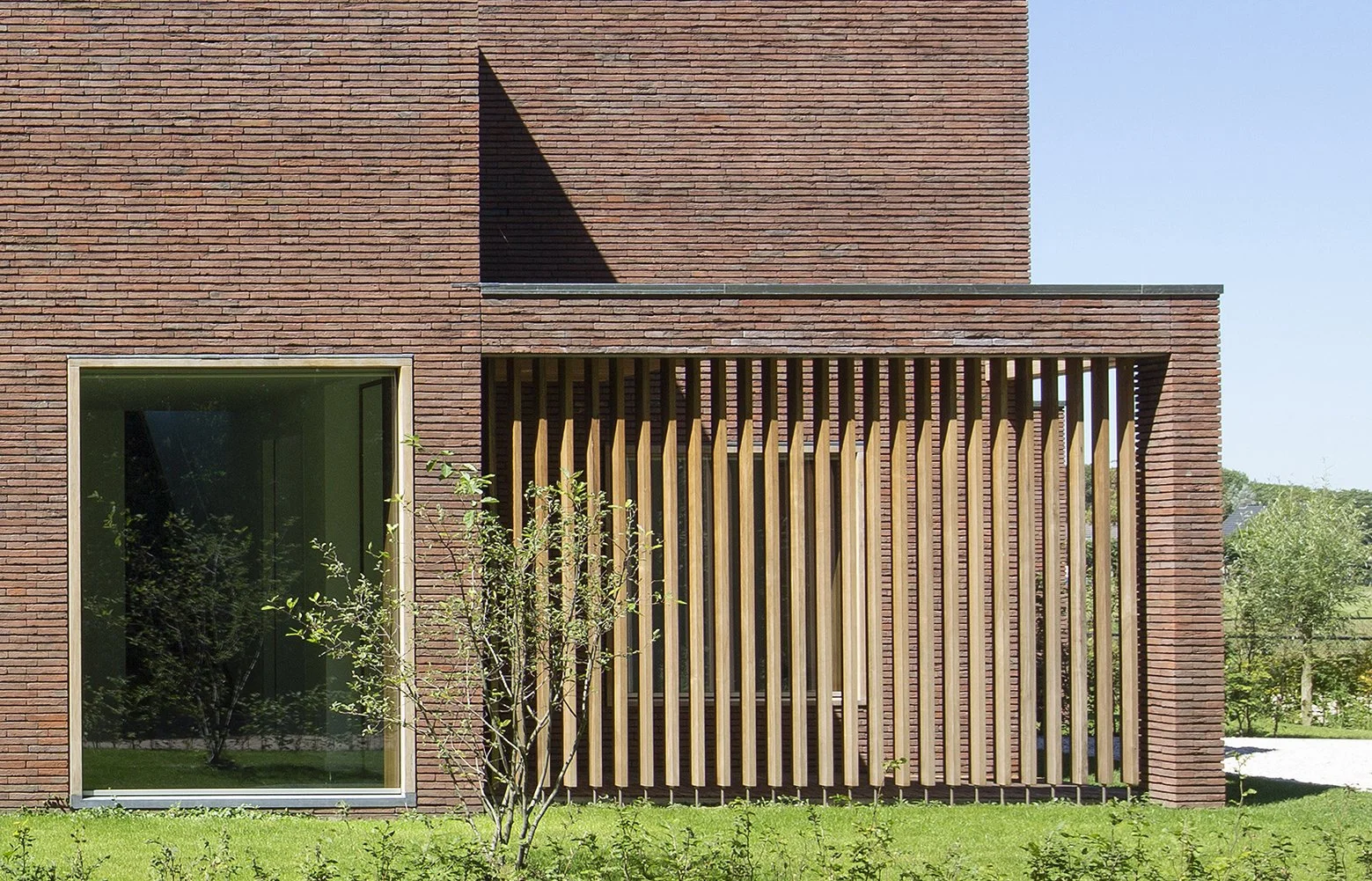Moderne gebouwmuur met grote raam en houten gevel, omgeven door groen gras en bomen.