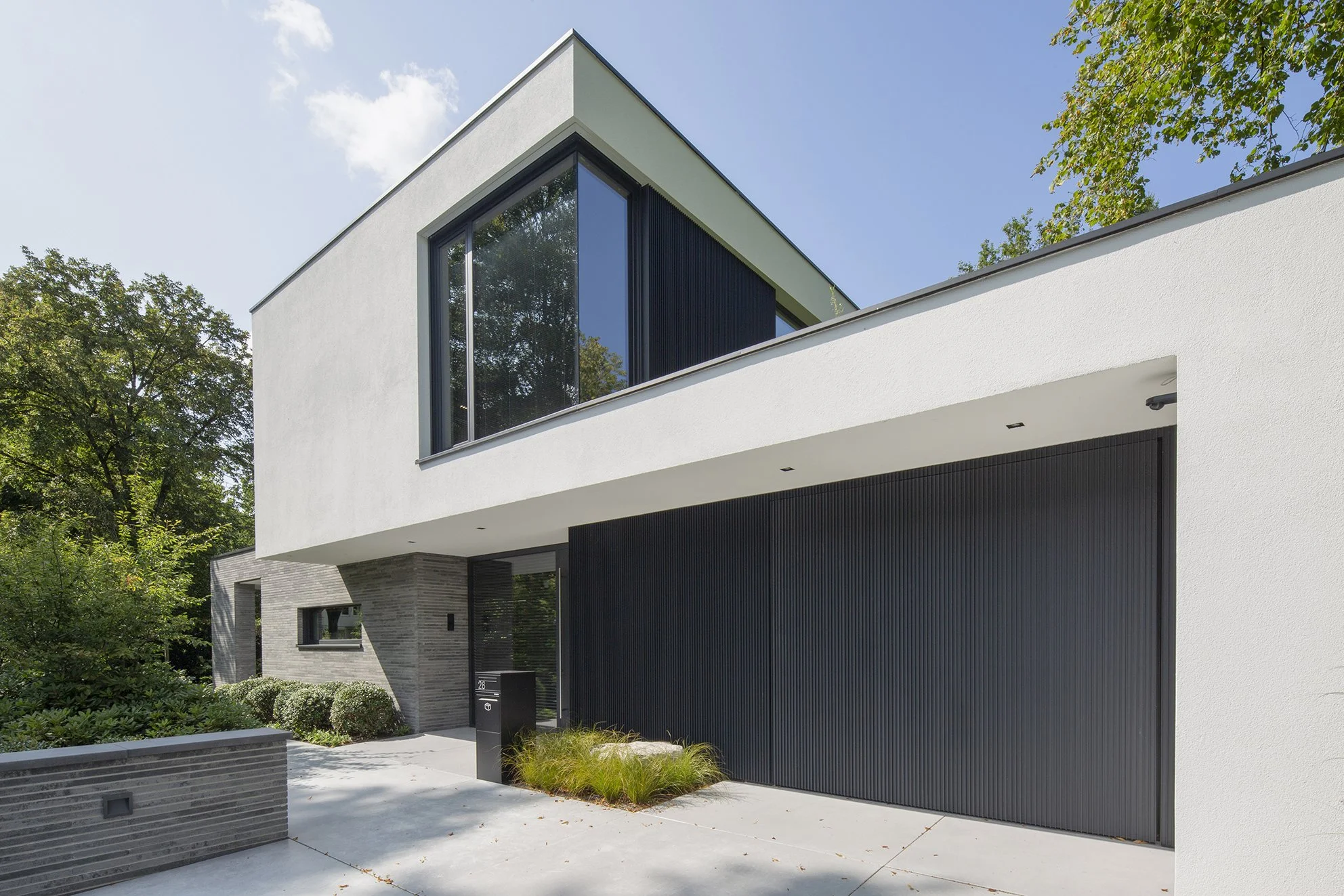 Moderne witte en zwarte woning met grote ramen, een balkon, omgeven door groen planten en bomen, onder een blauwe hemel.
