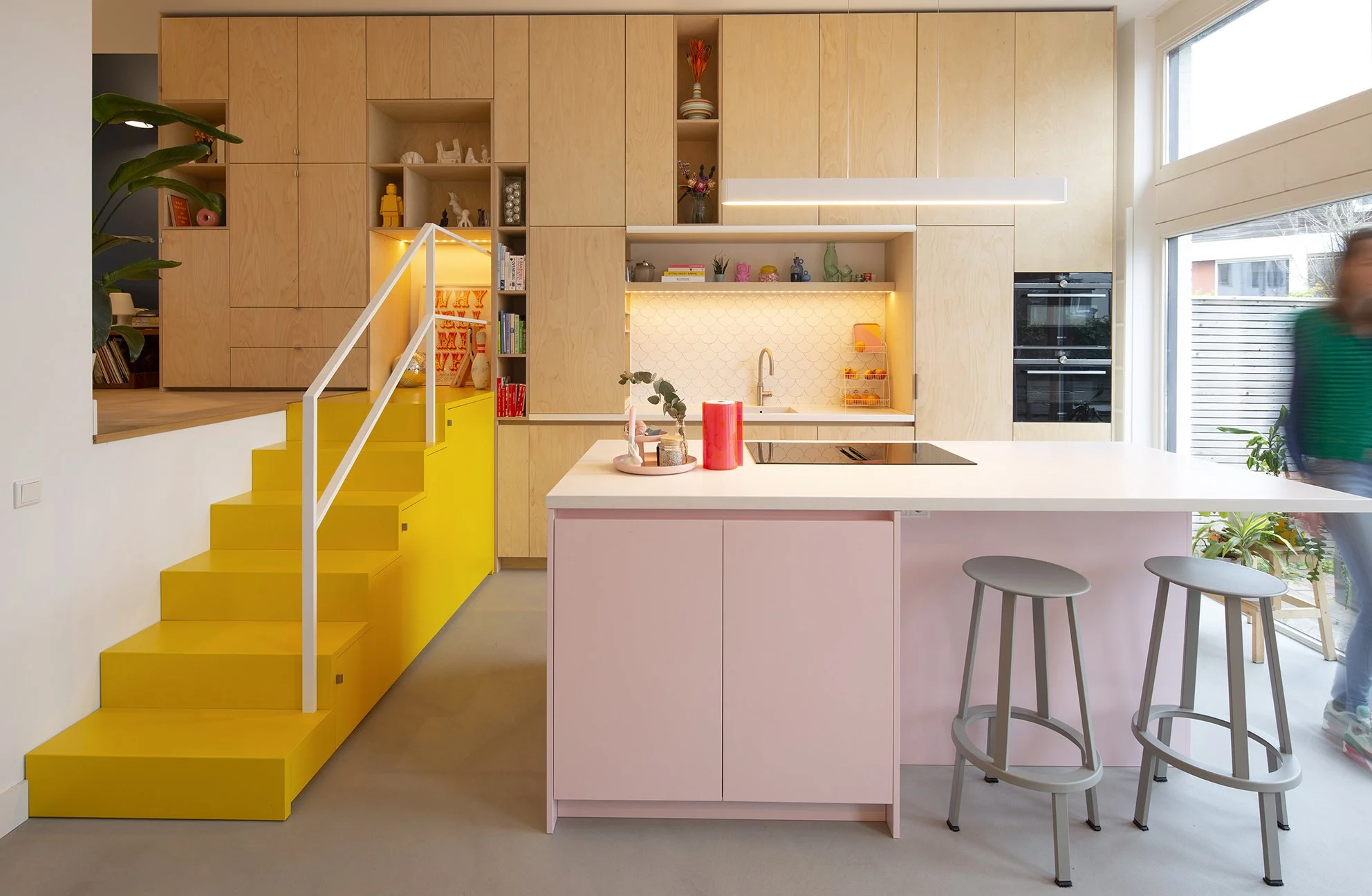 Moderne keuken met houten kasten en een witte eettafel met twee krukken, grote ramen die natuurlijk licht binnenlaten, en een trap met gele treden.