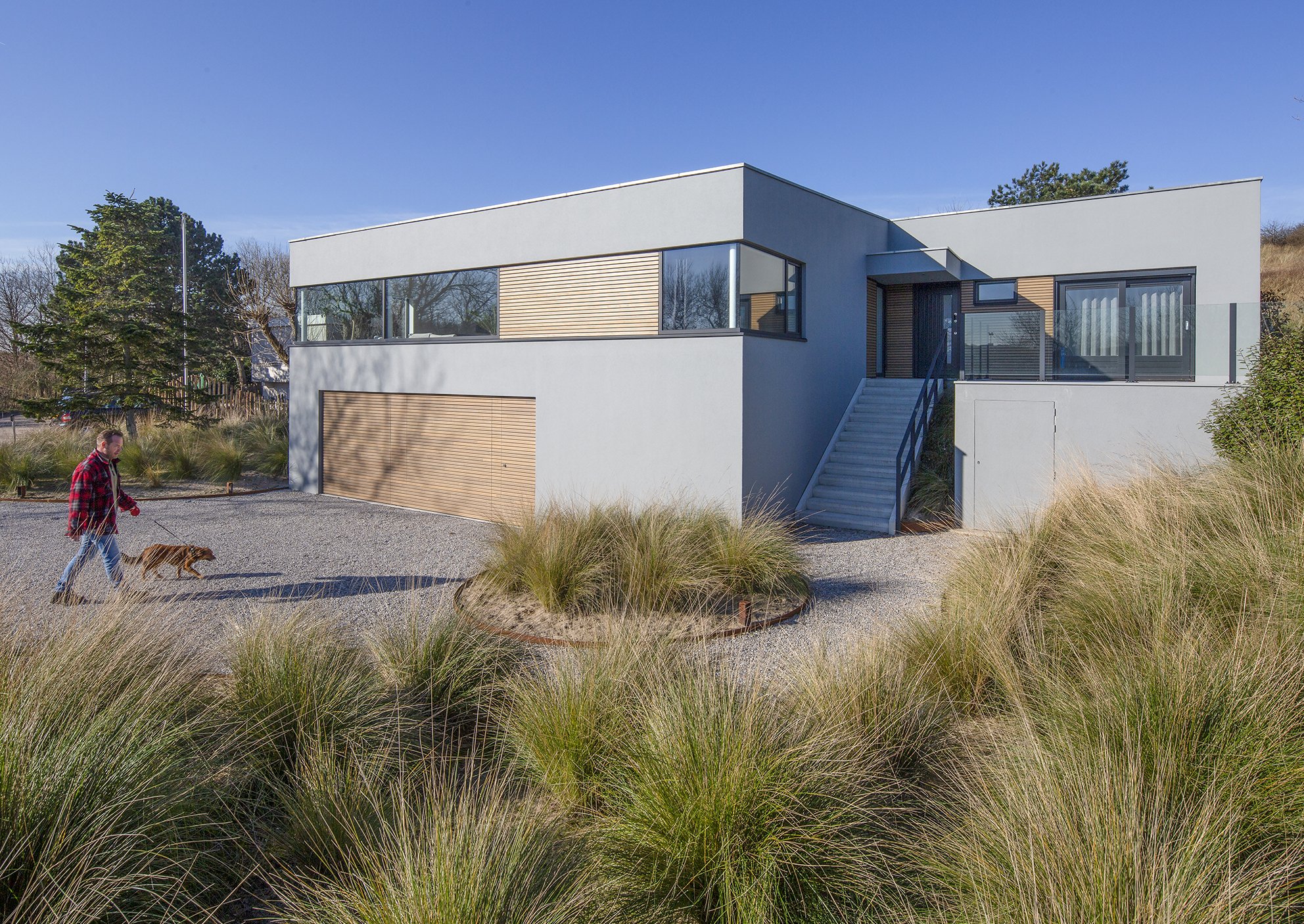 Moderne witte woning met grote ramen, houten details, trap en tuin met gras en struiken, onder een heldere blauwe lucht.