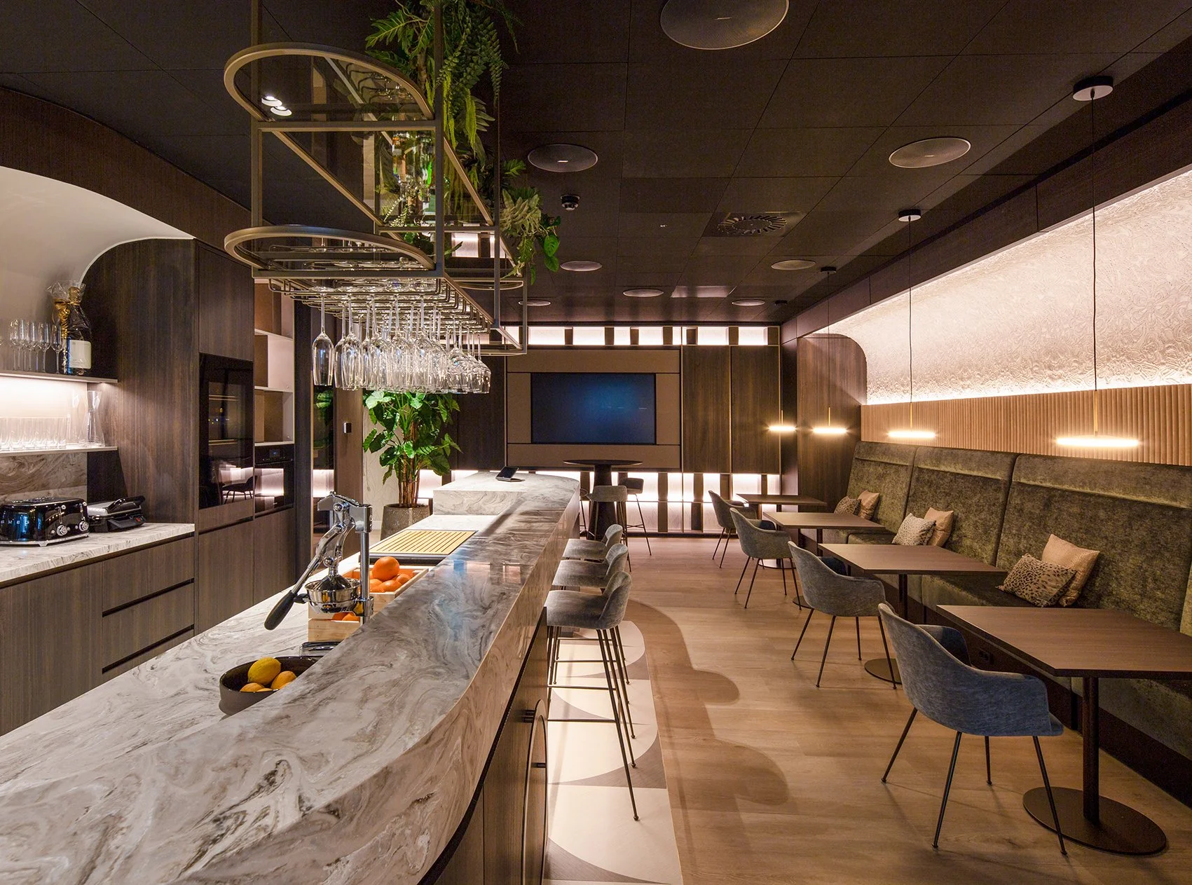 Moderne bar of lounge met een marmeren bar counter, barkrukken en een muur met een ontspannende inrichting. Verlichte plafondlampen hangen boven de tafel. Er is een grote tv aan de muur en groene planten voor decoratie.