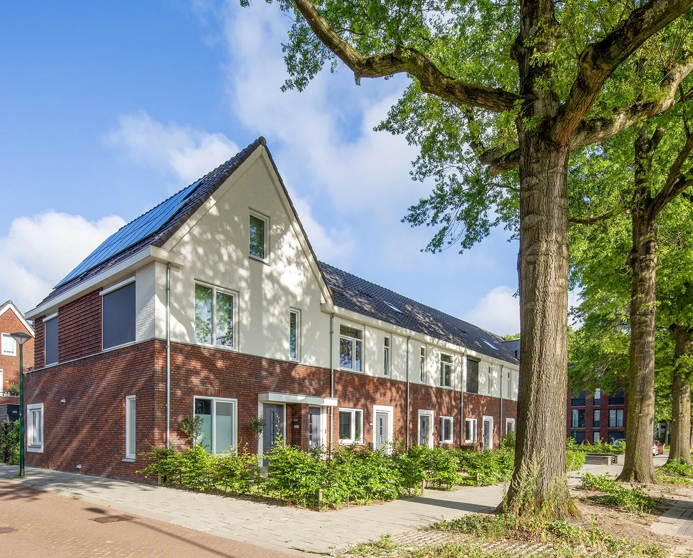 Moderne, witte en rode bakstenen woning met zonnepanelen op het dak, omringd door groene bomen en een voetpad in een woonwijk onder een blauwe lucht gefotografeerd door architectuurfotograaf John van Groenedaal.