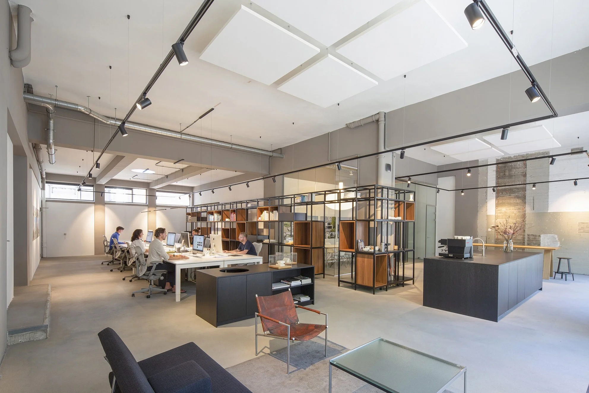Open kantoorruimte met mensen die aan computers werken, boekenplanken, zolderramen en moderne verlichtingsarmaturen.