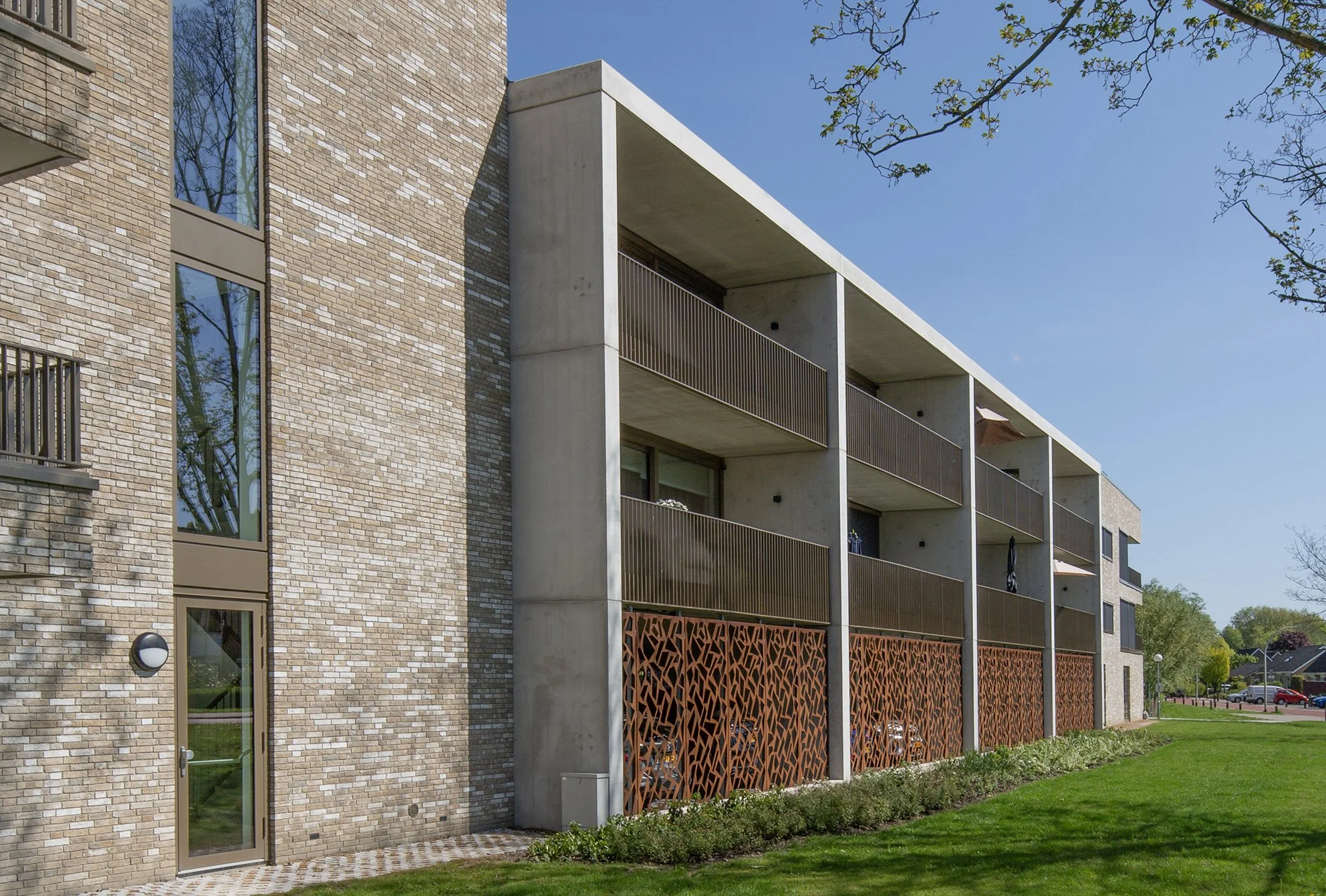 Moderne appartementencomplex met grote raampartijen en een balkon met houten reling, omgeven door groene tuin en bomen, onder een heldere blauwe hemel.