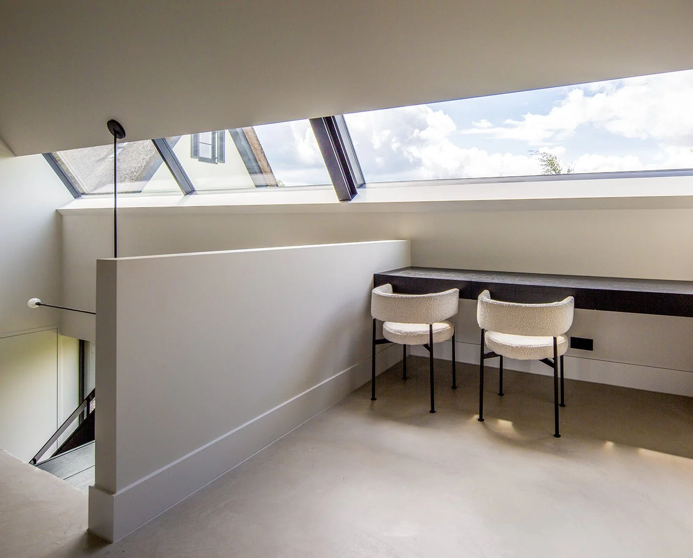 Binnenruimte met een schuine plafond met grote dakramen, twee witte stoelen naast een zwart werkblad onder het raam, en een trap aan de linkerkant.