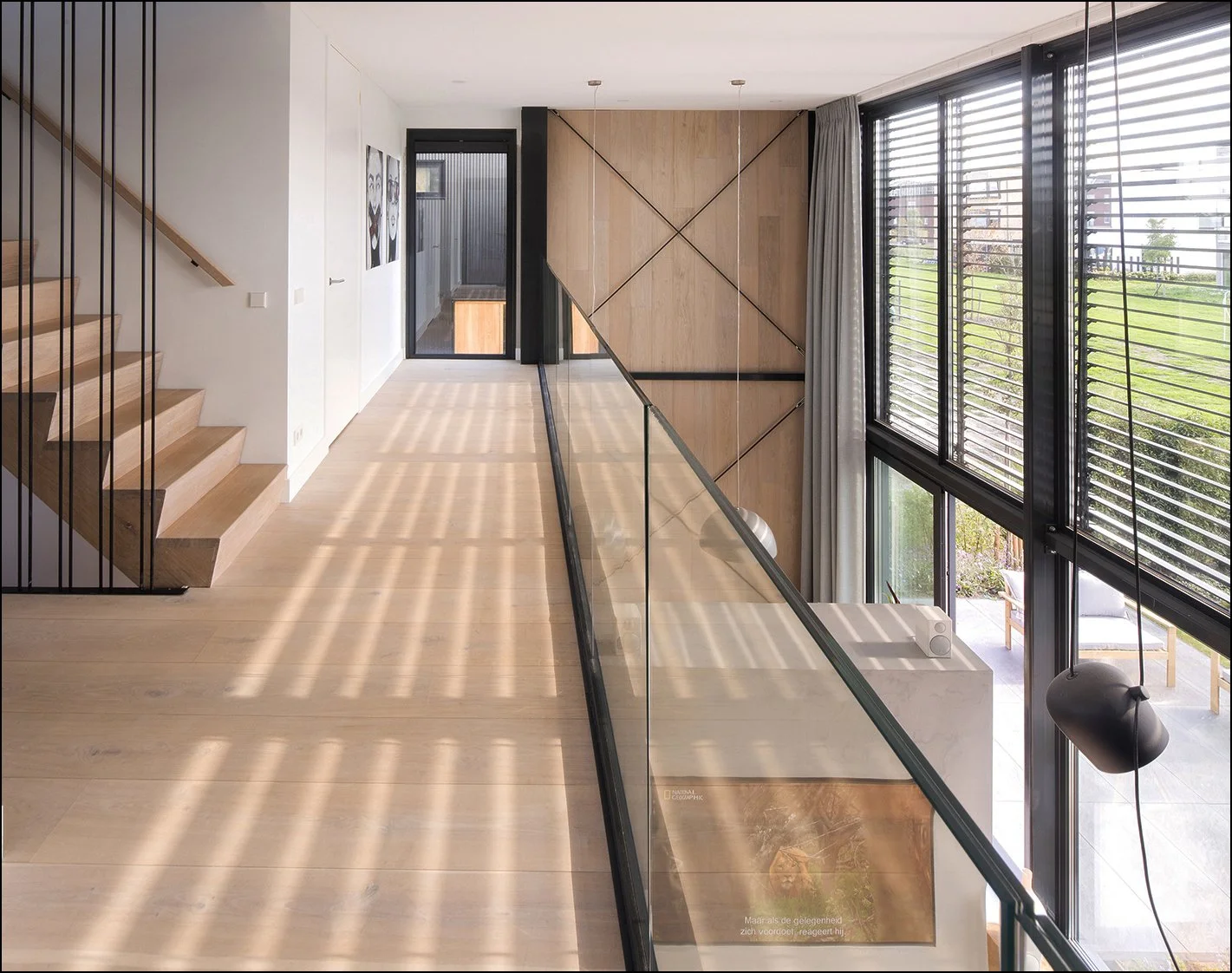 Binnenkant van een moderne woning met houten en glazen elementen, trappen, grote ramen met shutters en een zithoek buiten.