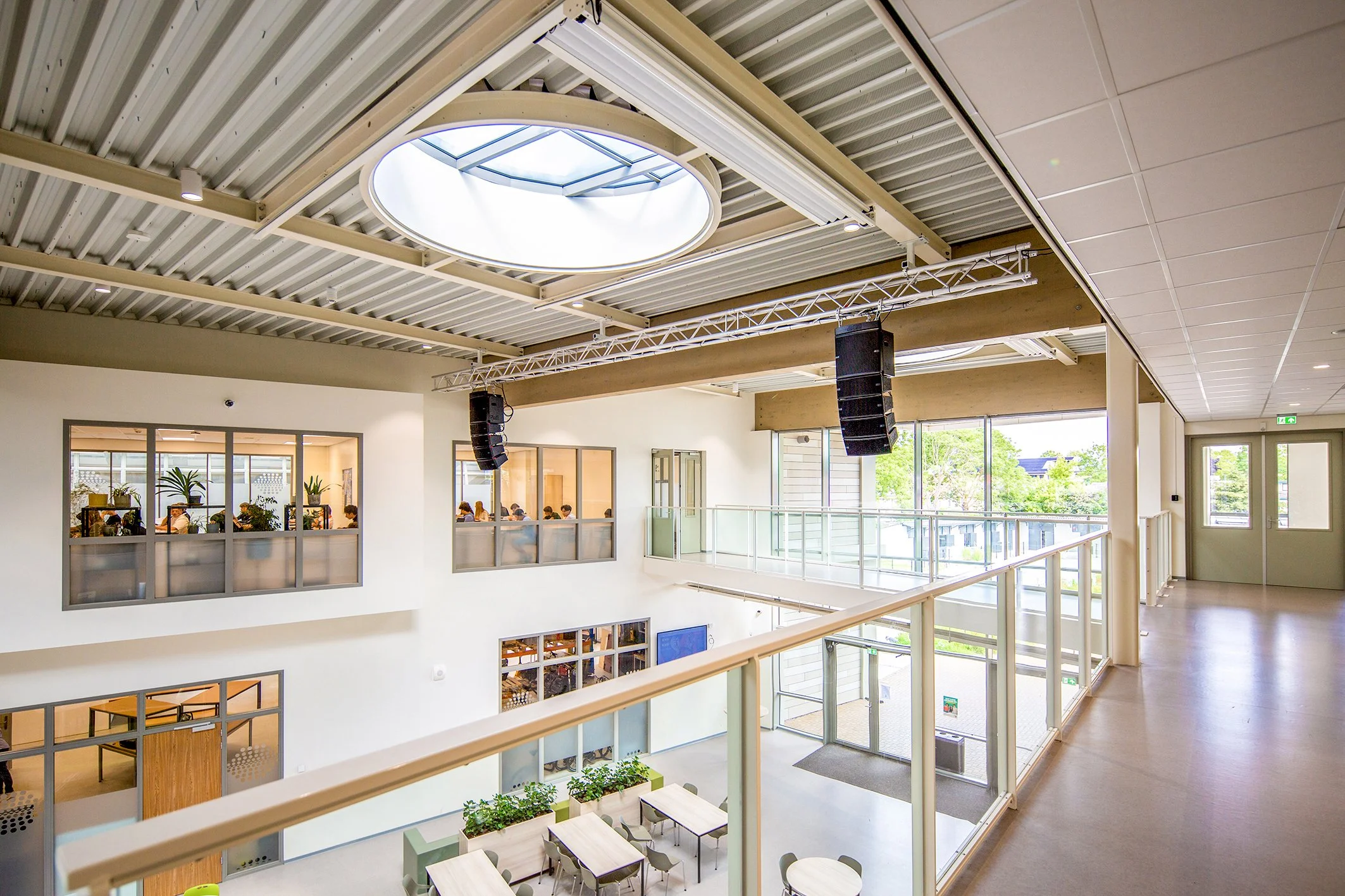Binnenruimte van een modern gebouw met meerdere verdiepingen, grote ramen en een open inrichting. Op de onderste verdieping zitten mensen in een ruimte met veel planten en tafels, terwijl de bovenverdieping een balkonsysteem heeft en een lichtkoepel 