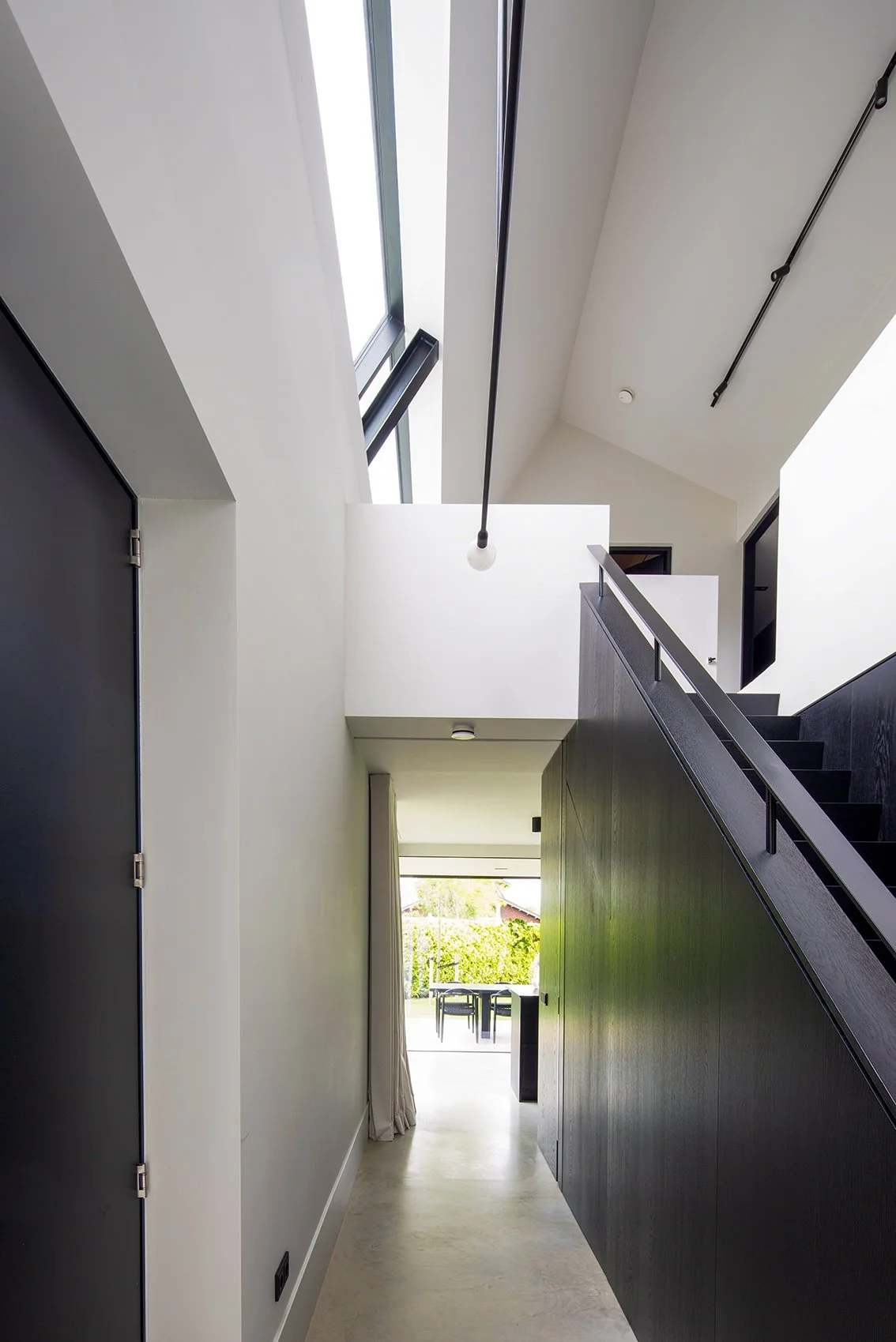 Binnenkant van een modern huis met een hal, trap en grote ramen die natuurlijk licht binnenlaten, met uitzicht op een tuin.