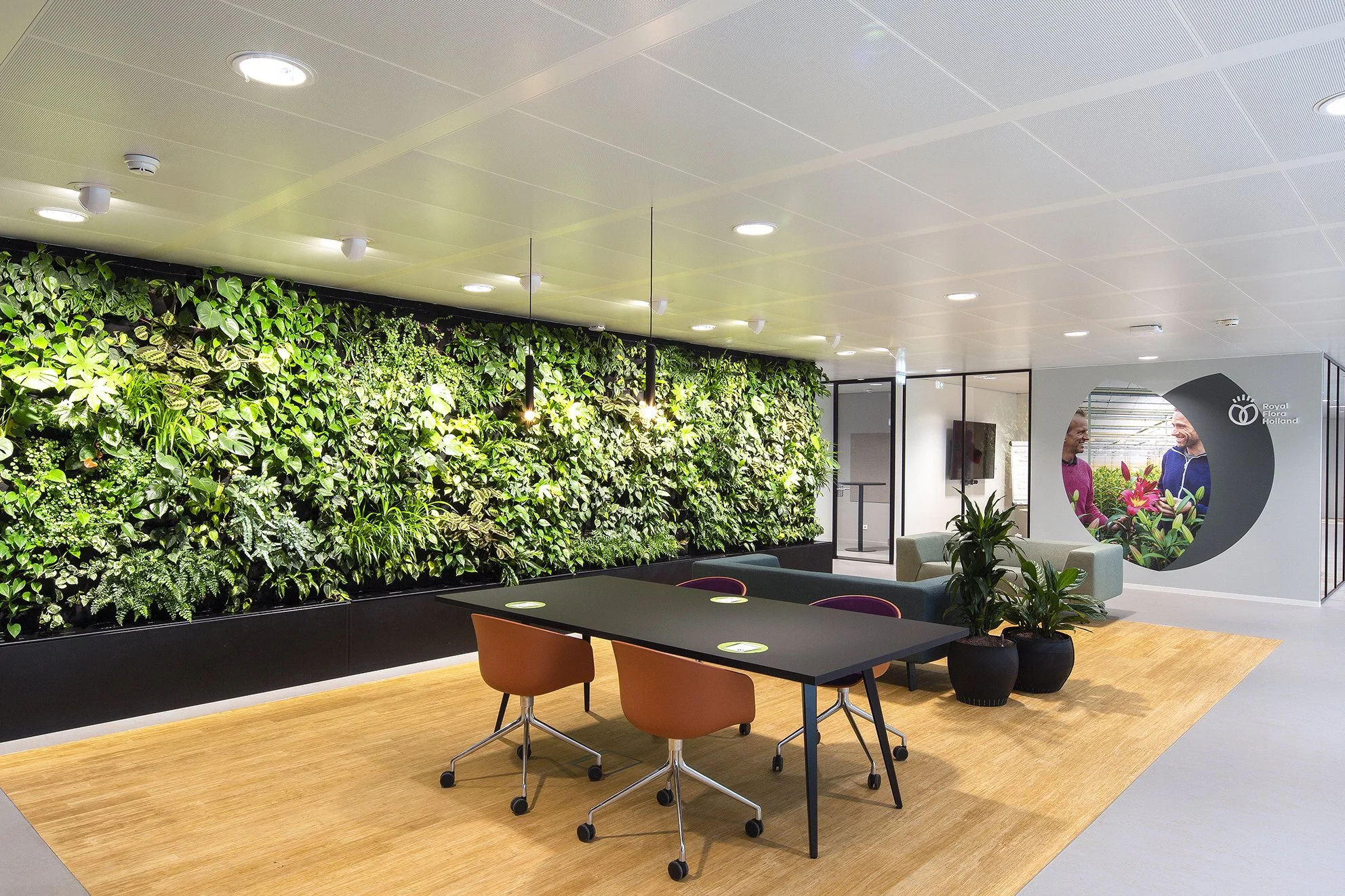 Binnenruimte met een groene muur van planten, een zwarte tafel met oranje stoelen, een beige bank, enkele planten en een muur met een logo van Royal Flora Holland, je zitruimte met mensen die lachen.