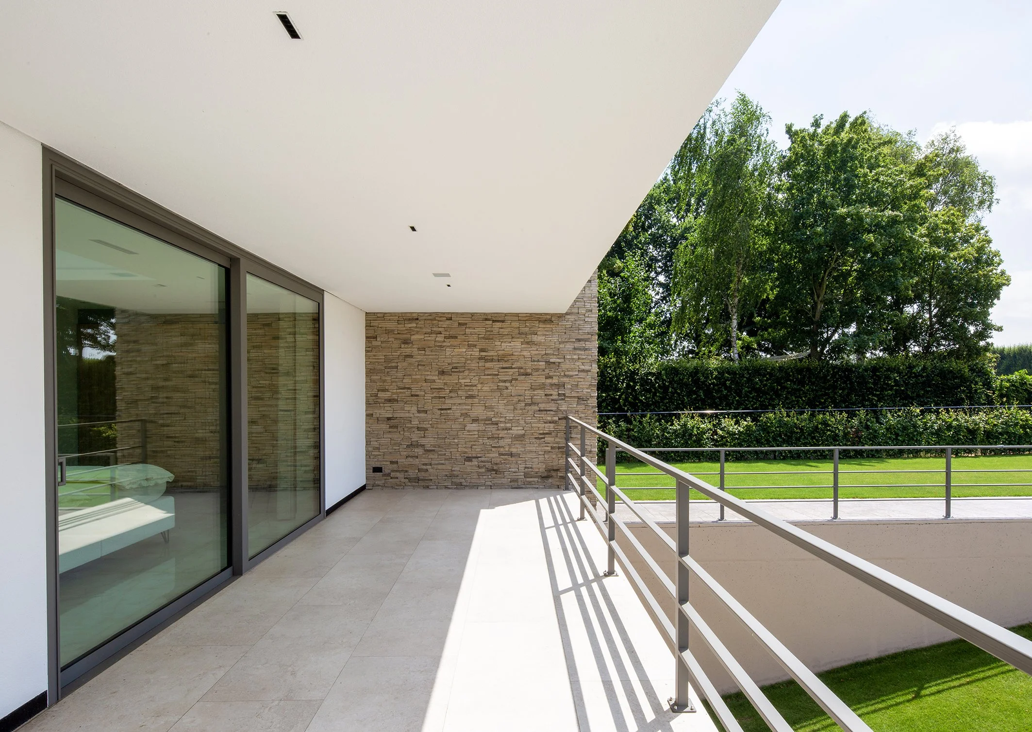 Een modern balkon met glazen deuren, een muur van bakstenen en een groene tuin, met een witte plafond en een metalen railing.