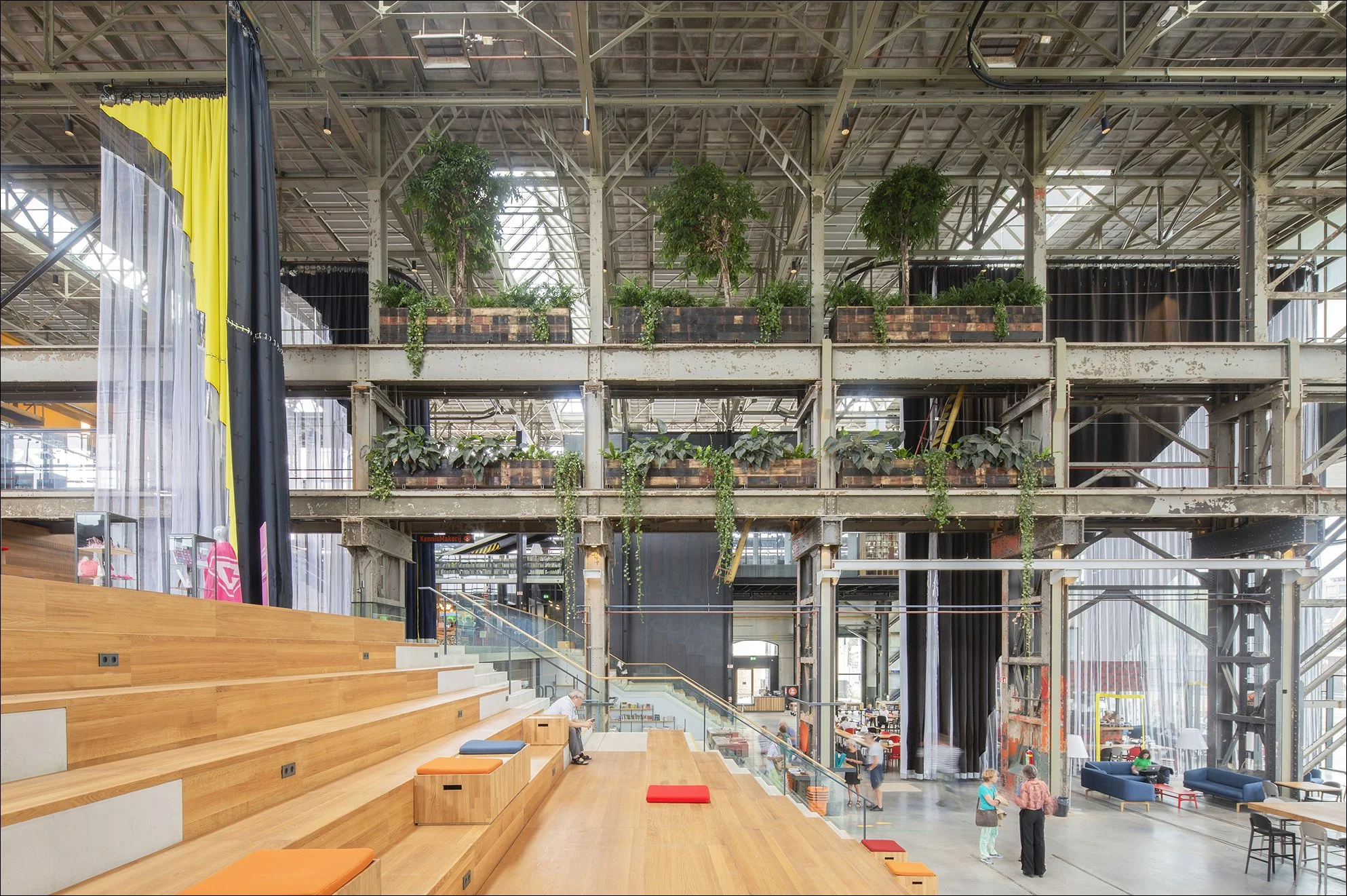 Binnenkant van een modern gebouw met groene planten op een bovenverdieping, houten zittribune en mensen die zich bevinden op de begane grond en bovenverdiepingen.