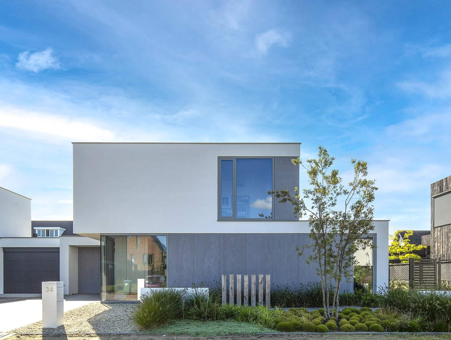 Moderne witte en grijze woning met grote ramen en een tuin met een kleine boom en grassen onder een blauwe lucht.