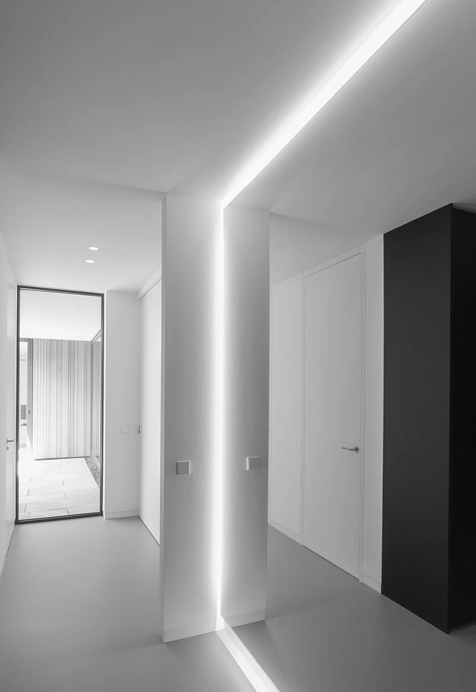 Moderne, minimalistische entree met witte muren, donker accentmuur, plafondverlichting en glazen deur naar buiten.