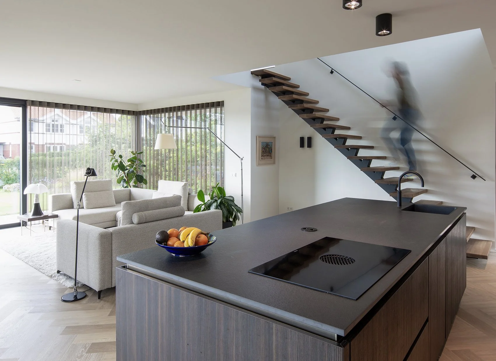 Moderne woonkamer en keuken met open trap, een bank, planten, grote ramen met verduisteringsgordijnen, fruitschaal op het eiland, en wanddecoratie.