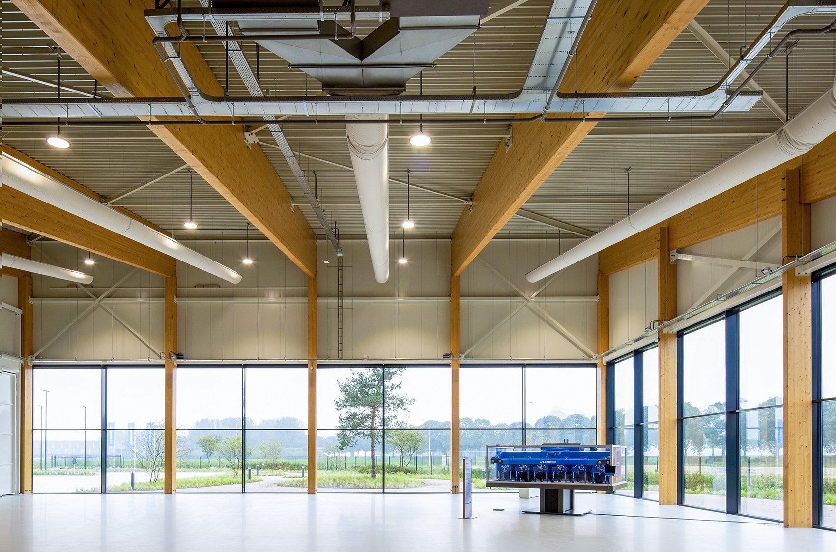 Interieur van een moderne grote hal met grote ramen, houten balken, ventilatiesystemen en verlichting, uitzicht op buiten met bomen en landschap.