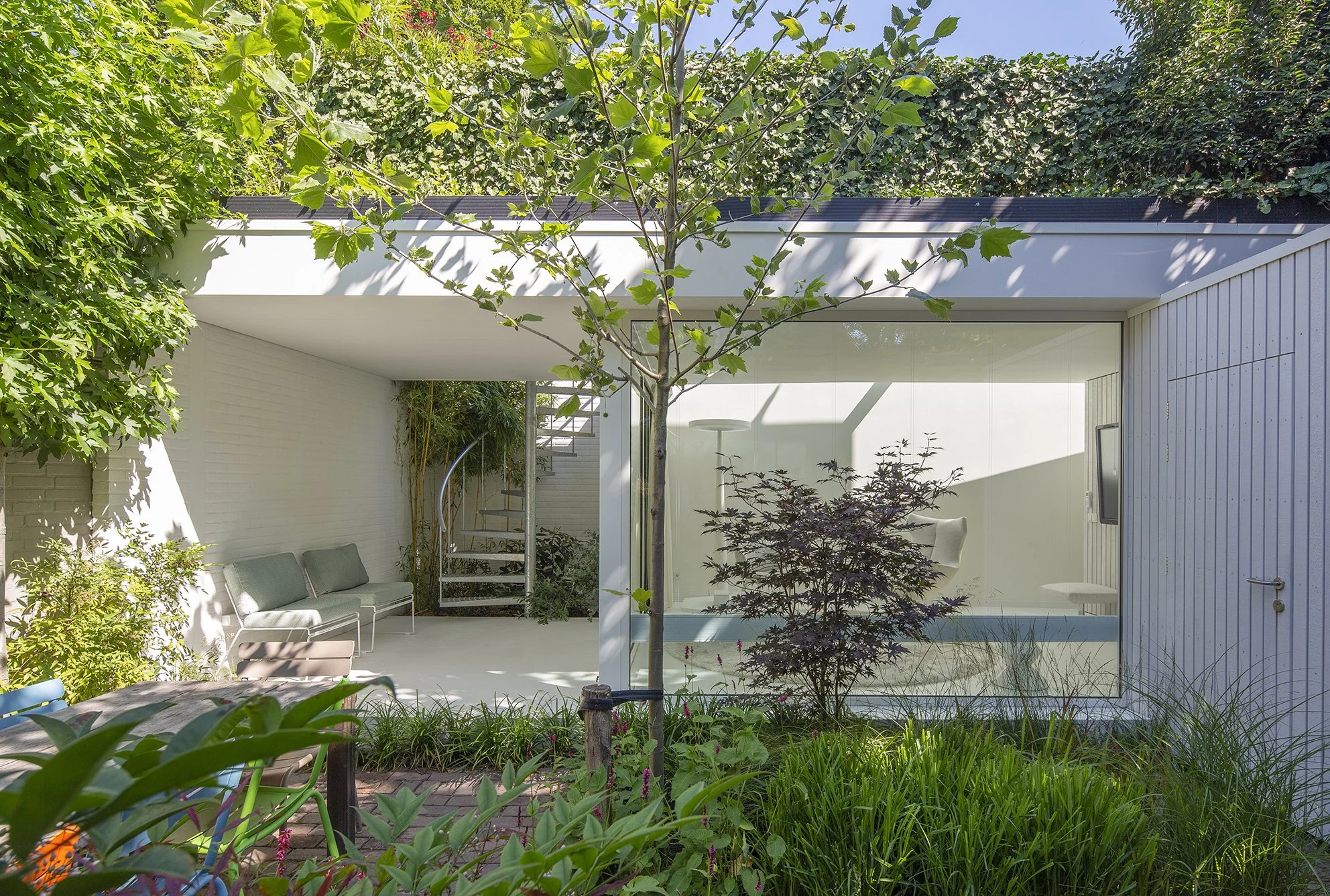 Een modern huis met grote ramen, omgeven door groene planten en bomen. Er is een tuin met zitbank en tuinmeubilair, en een trap die naar het bovenste niveau leidt.
